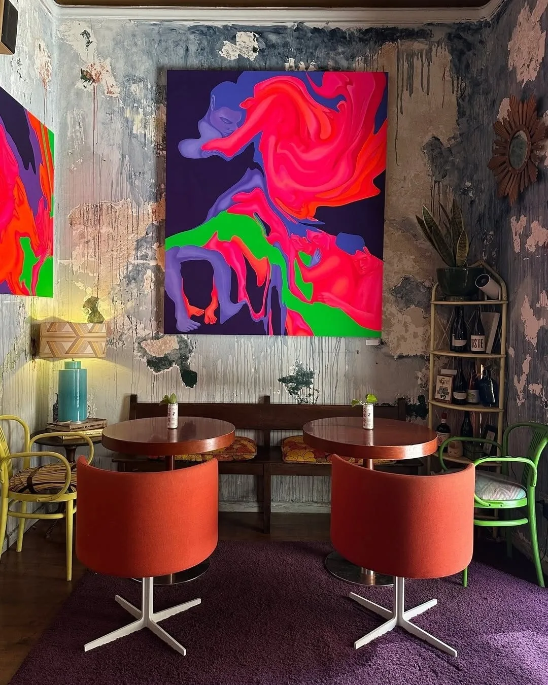 A colourful bar corner with orange chairs, small round tables and a large painting on the wall