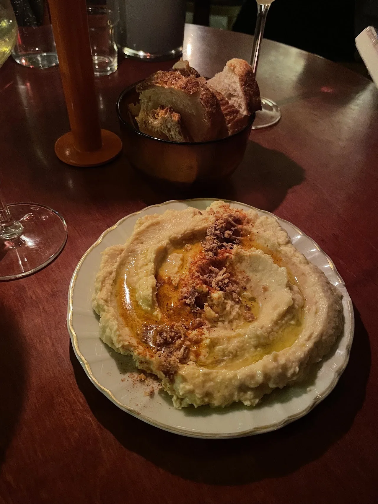 A plate of hummus with a glass of red wine and a bread basket on a dark table