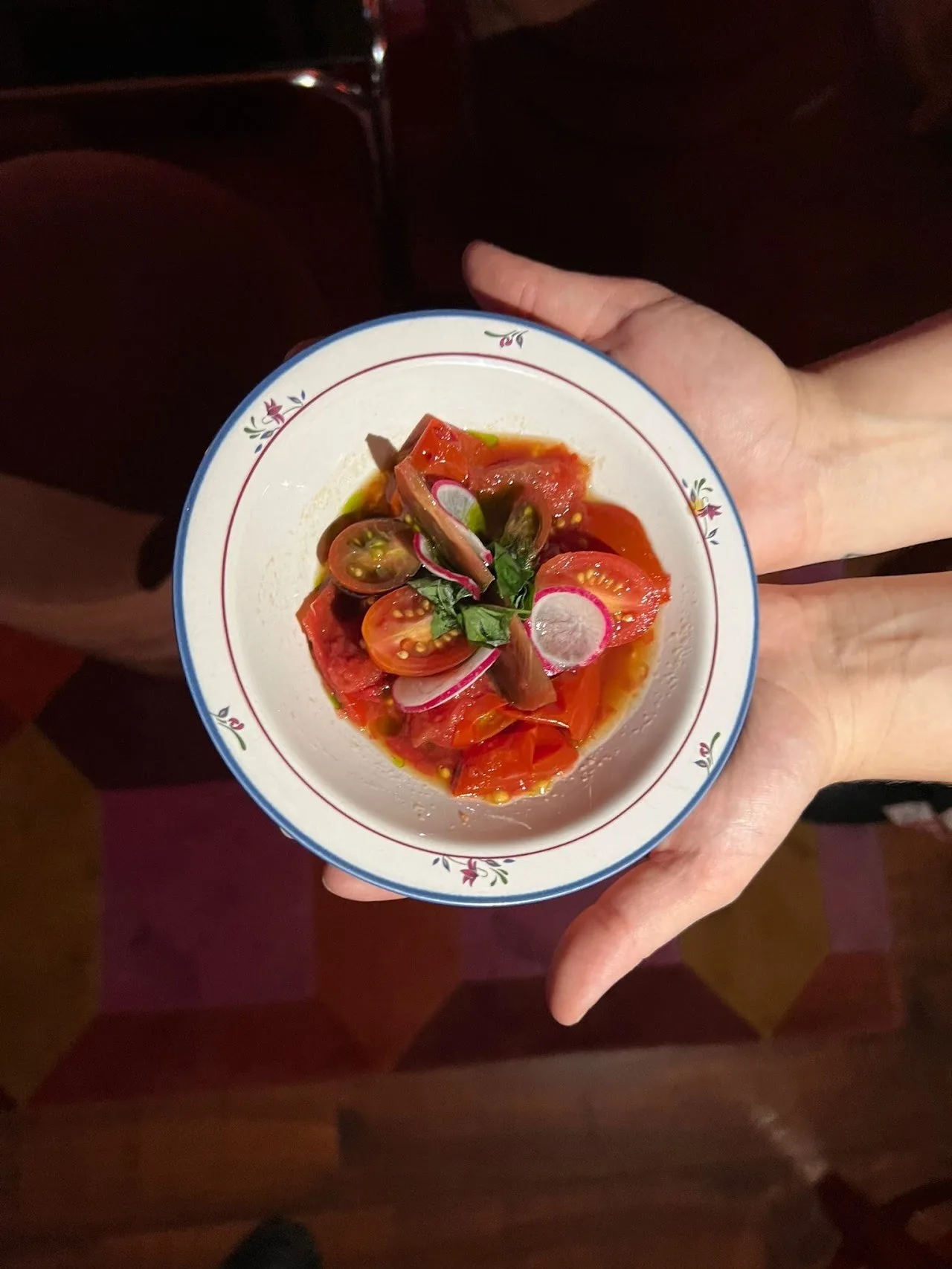 A hand holding a white bowl of tomato-based dish over a patterned floor