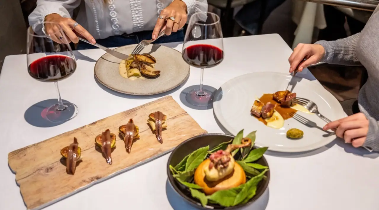 Table spread of dishes including anchovy-and-fig toasts on a wooden board, a foie and mushroom plate, seared meat in sauce and a bowl garnished with bay leaves, with two glasses of red wine