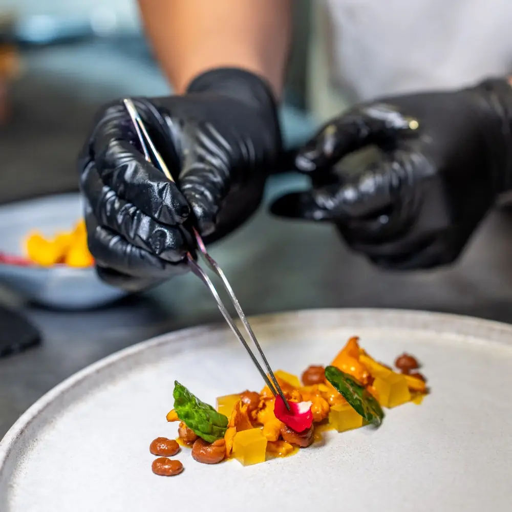 Black-gloved hands placing a flower petal with tweezers on a plated dish of chanterelles, beans and diced mango