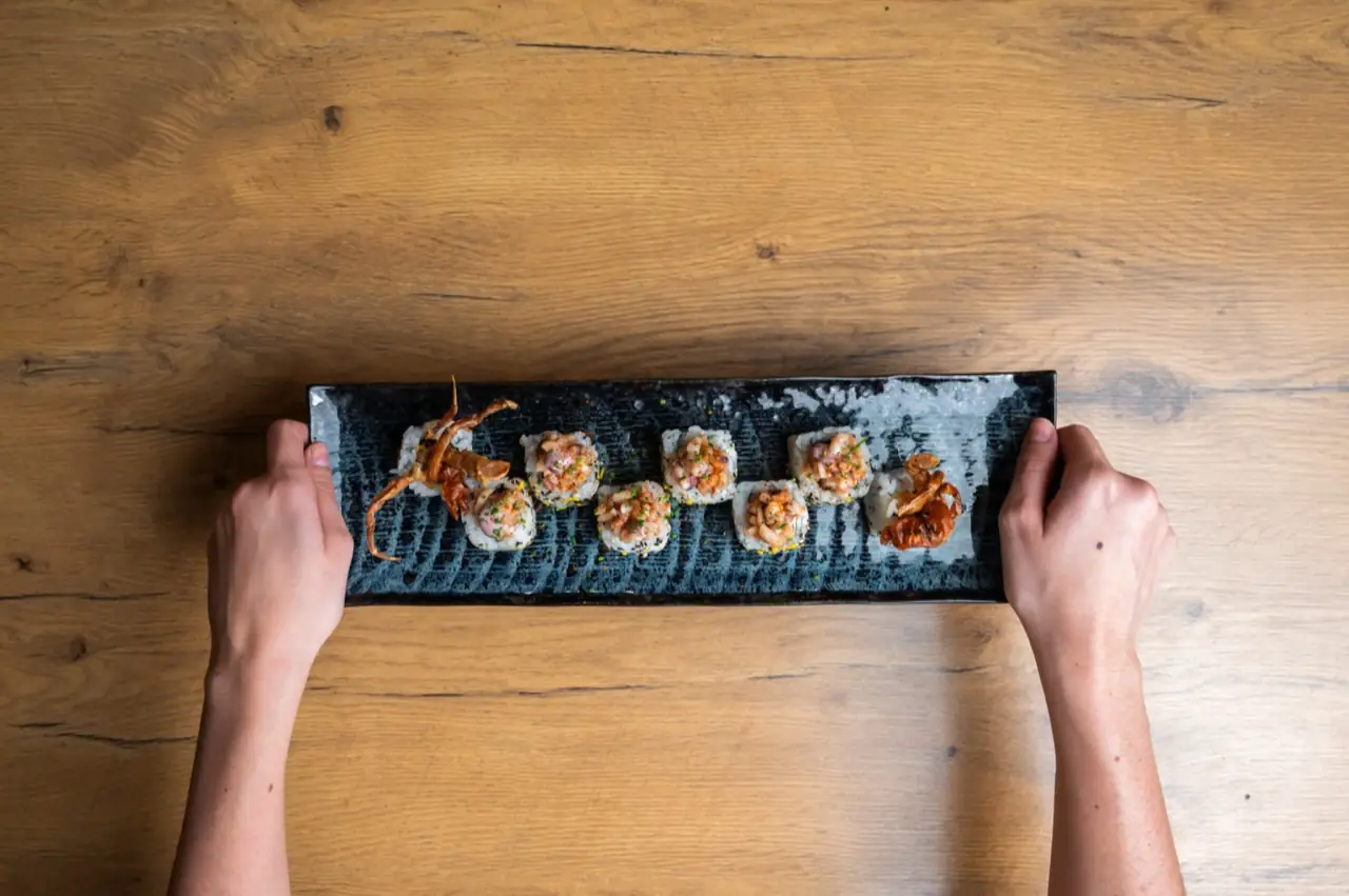 Row of sushi rolls topped with chopped shrimp on a long black slate plate held by two hands over a wooden table
