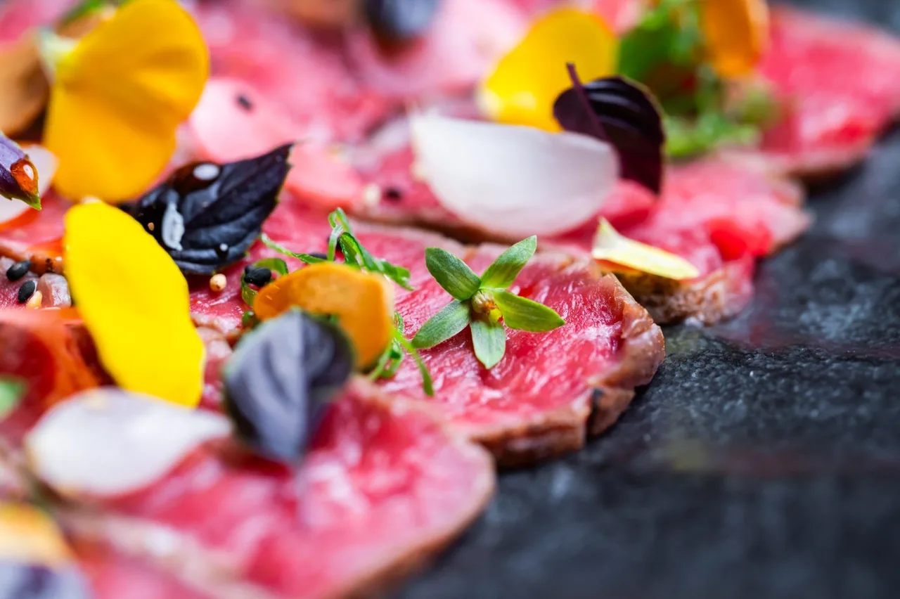 Thin slices of raw red meat garnished with edible flowers and herbs on a dark slate