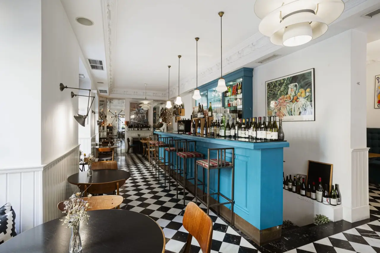 Interior with a bright turquoise bar counter lined with wine bottles, metal barstools, black-and-white checkerboard floor, pendant lamps and a fruit still-life painting on the white wall