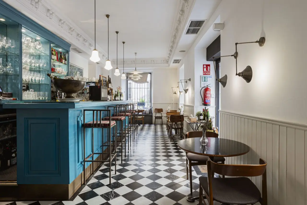 View along the turquoise bar with an illuminated glass cabinet of stemware, a silver ice bowl, espresso machine and wine bottles, brass wall sconces opposite and checkerboard floor
