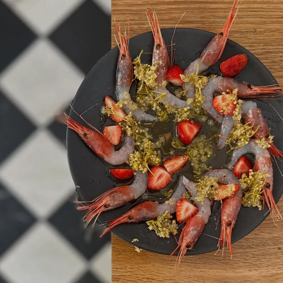 A dark round plate of red prawns arranged with sliced strawberries and green garnish