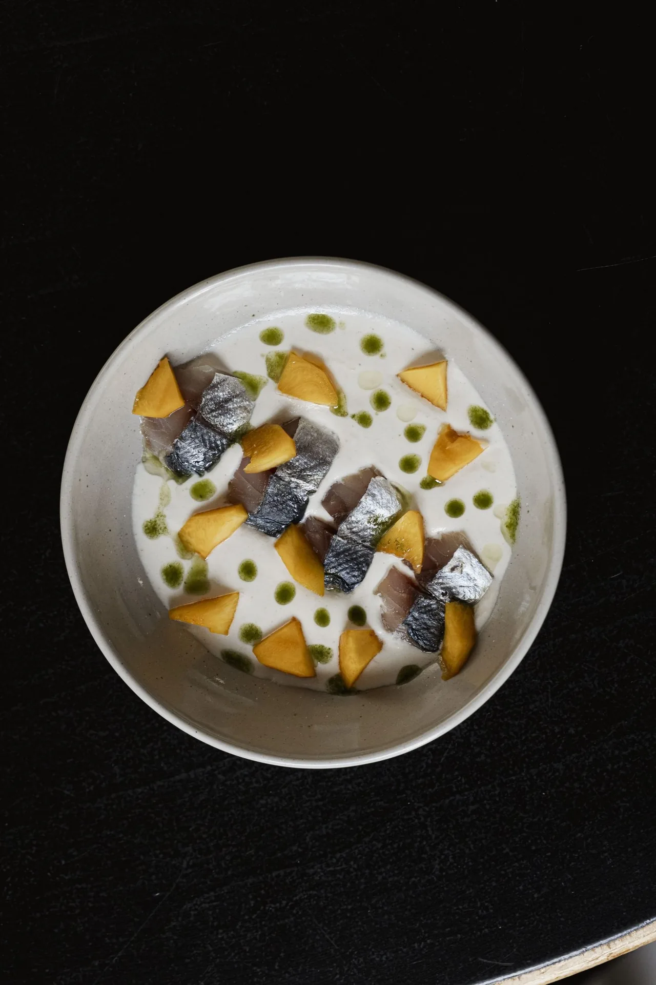 Overhead view of a round white bowl with sliced fish, orange pieces and green dots on a black table