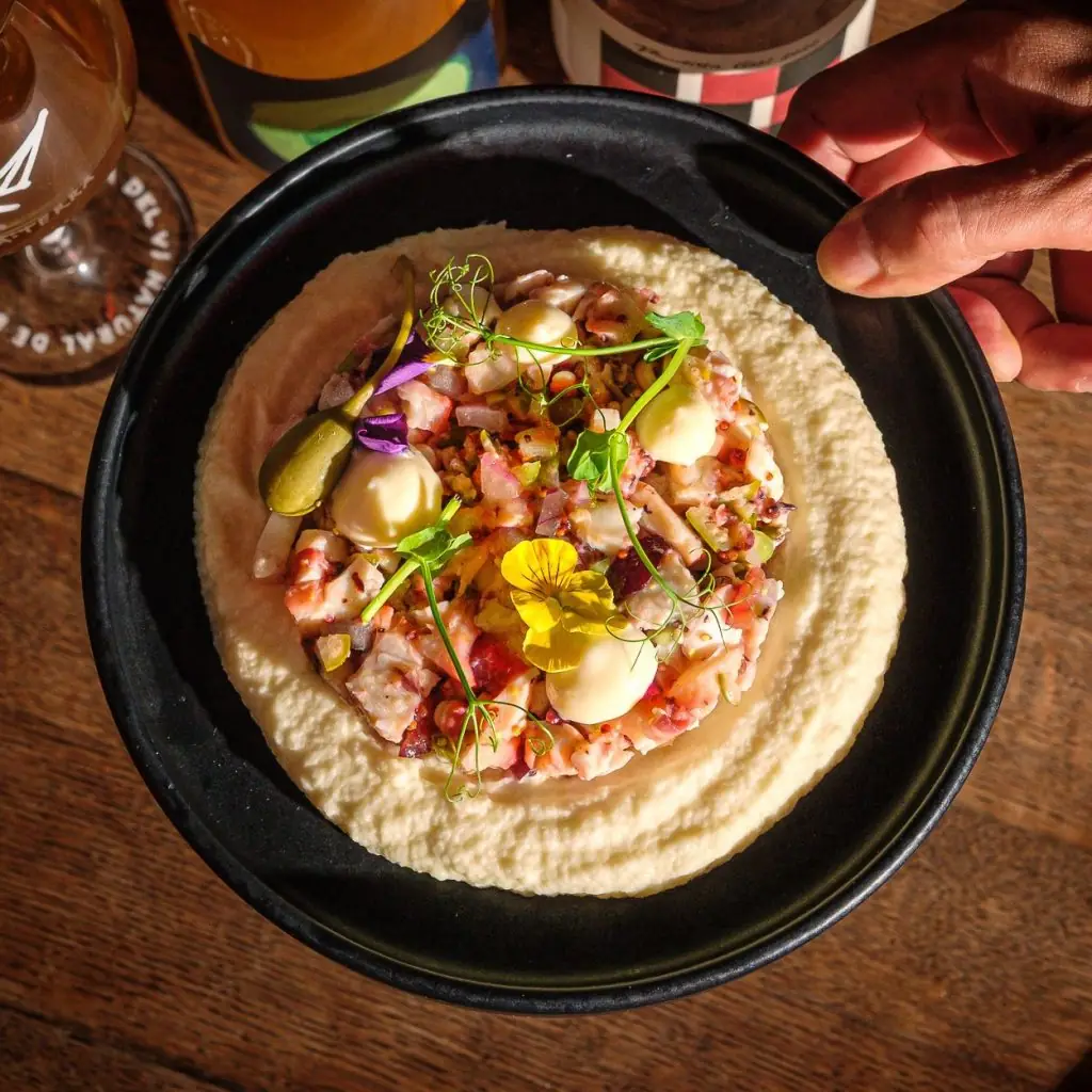 Octopus tartare over whipped potato with pickles and edible flowers on a black plate, Garage Bar