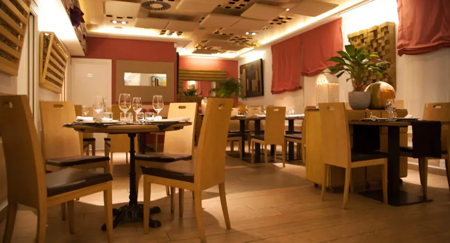Dining room with pale wood chairs and dark square tables laid with wine glasses and folded napkins, terracotta walls, red blinds and a potted palm under warm ceiling lighting