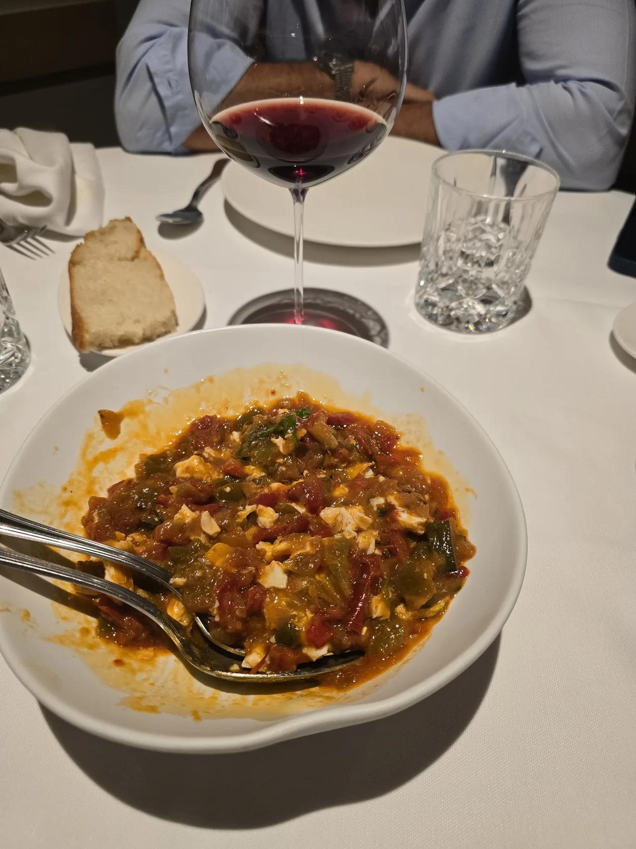 A white bowl of a tomato-based dish beside a glass of red wine