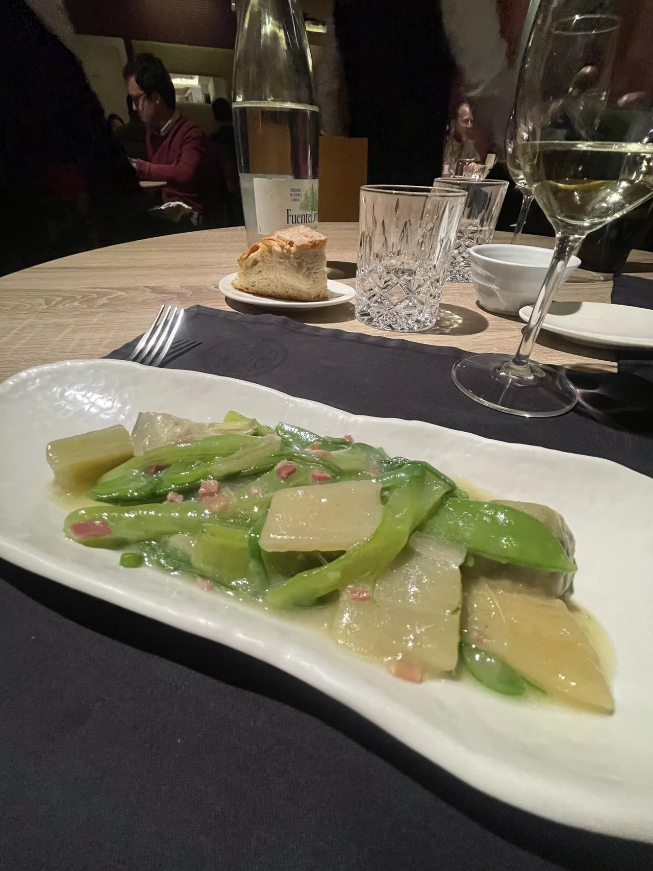 A plate of green vegetables beside a glass of white wine on a set table