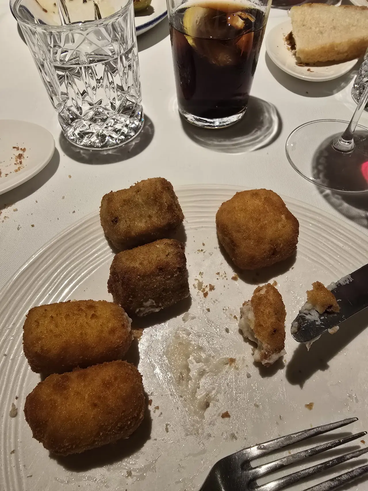 Square breaded croquetas on a white plate beside glasses at the table