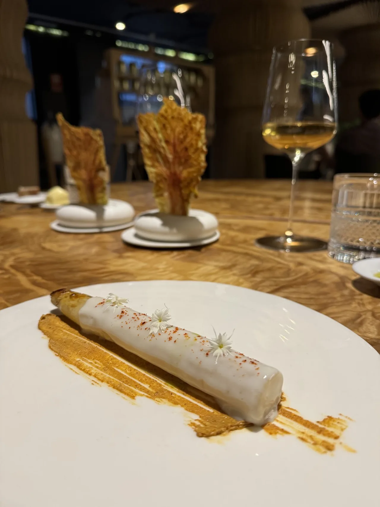 A white plate with a pale cylinder on golden sauce, on a wooden table with wine glasses and crisp garnishes