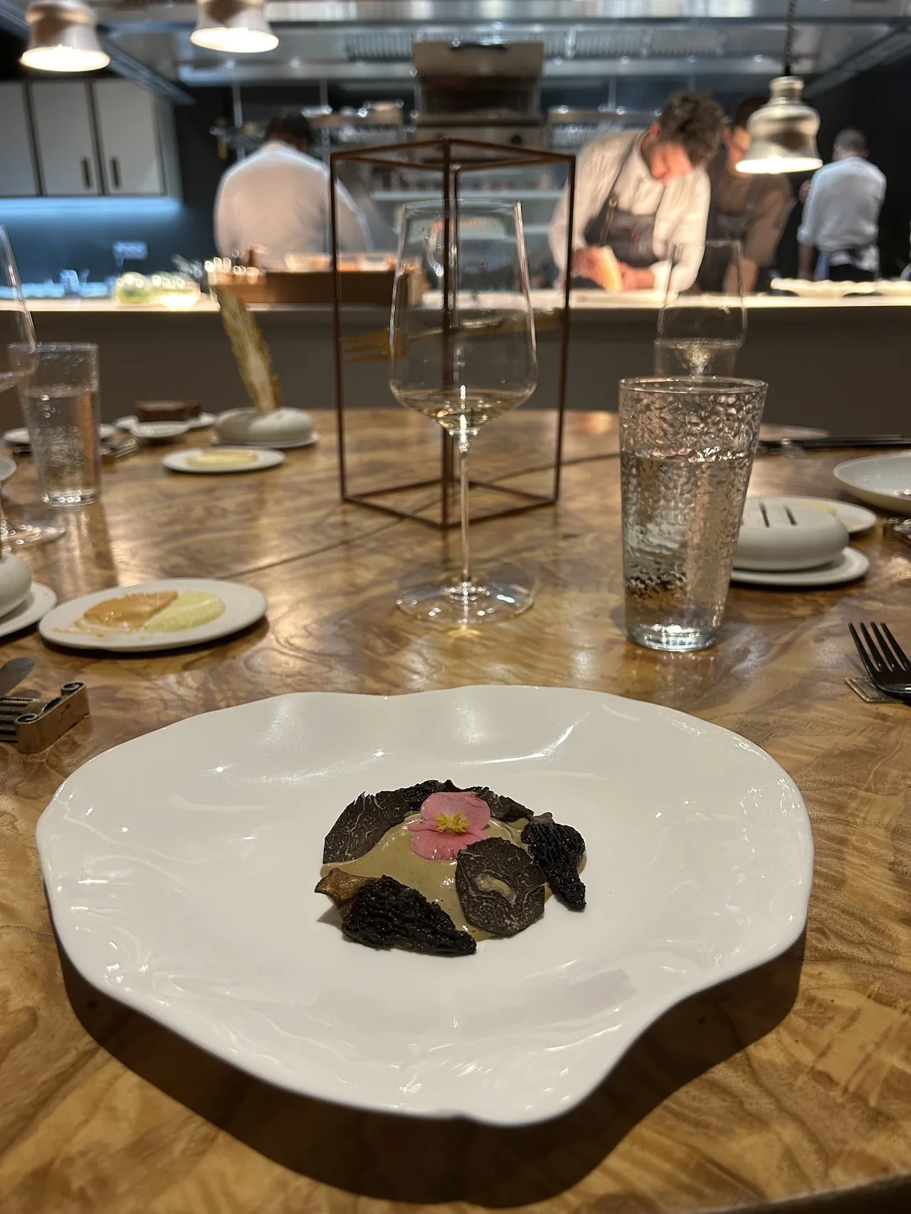 A white plate with a dark truffle-topped dish on a wooden table with glasses and an open kitchen behind
