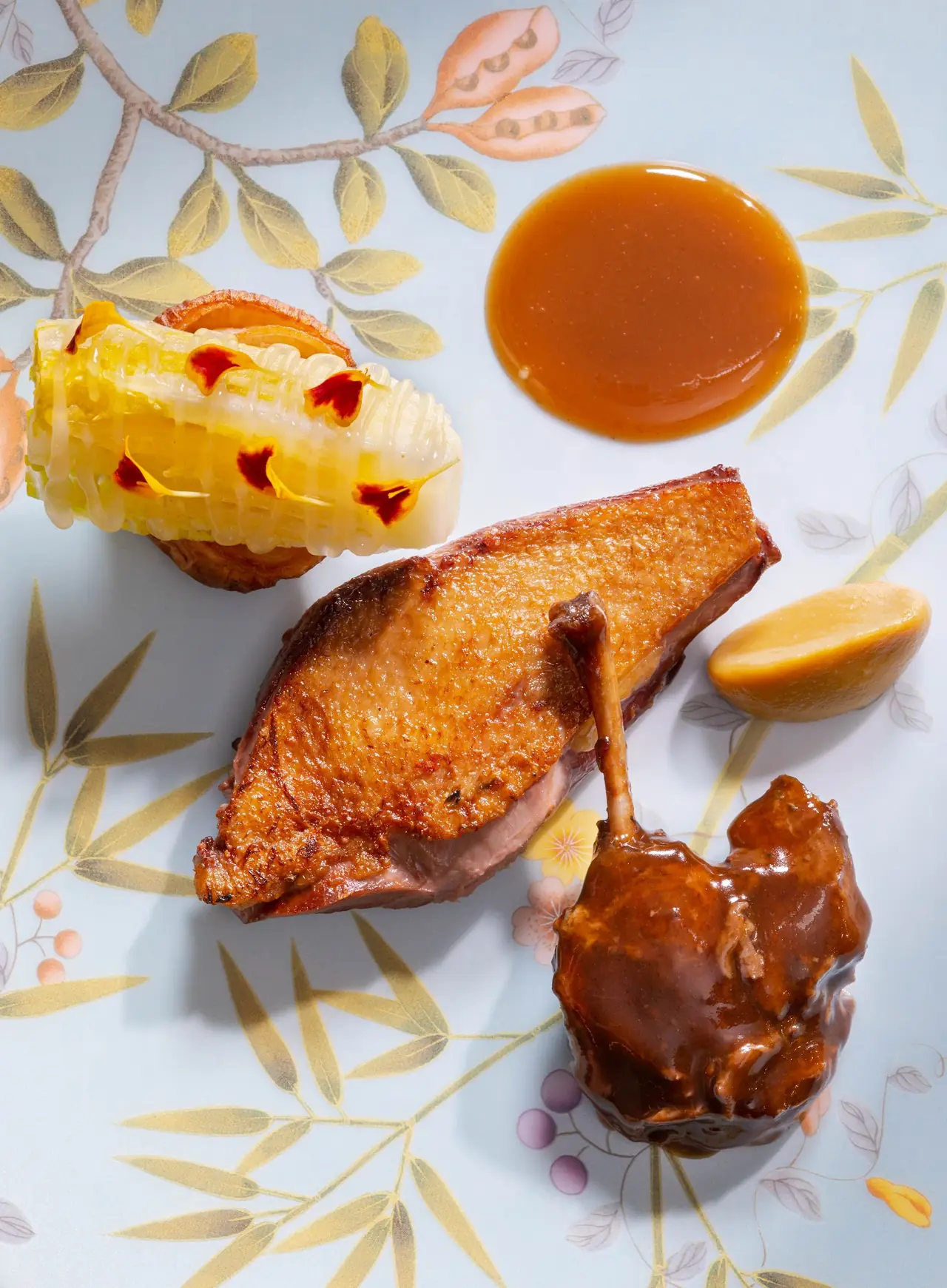 Roasted duck breast and confit leg with jus, glazed corn, and mango on a decorative floral-print plate