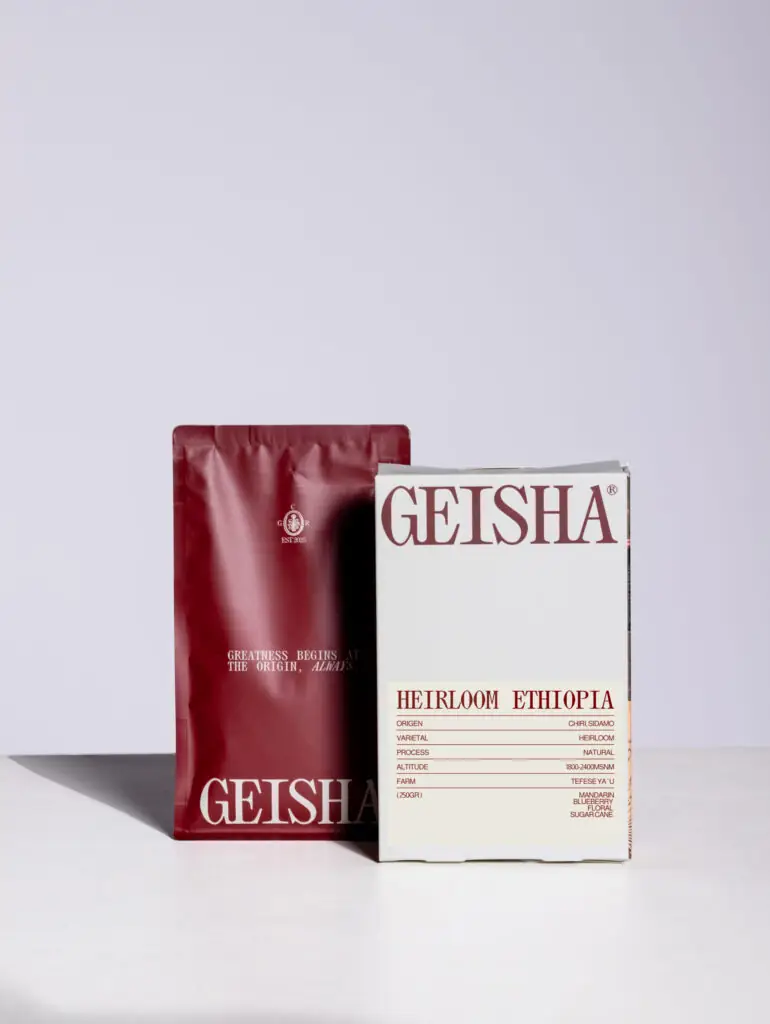 Composed retail product shot of Geisha specialty coffee: a maroon foil bag beside a cream box labelled 'Heirloom Ethiopia' with origin details, on a pale lilac backdrop