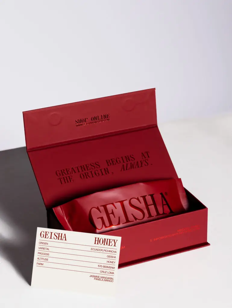 Open maroon Geisha gift box holding a foil coffee pouch, with a cream spec card reading 'Geisha Honey, Ecuador' on a lilac surface