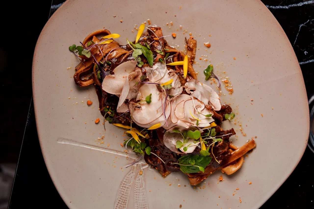 A ribbon pasta dish with a dark sauce topped with sliced raw mushrooms, herbs and yellow petals on a large pale plate