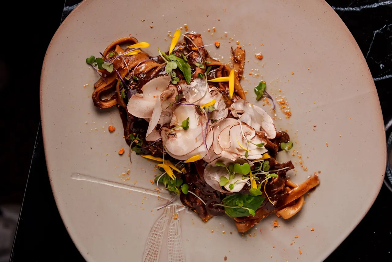 Plated pasta ribbons in dark sauce topped with sliced mushrooms, microgreens and edible flowers