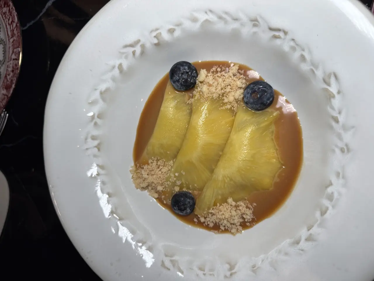 Plated dessert of three folded mango slices in caramel sauce with blueberries and crumble on a decorative white plate