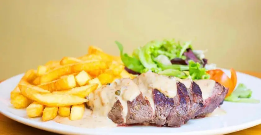 Sliced steak with peppercorn sauce, fries, and mixed salad on a white plate, Gilda