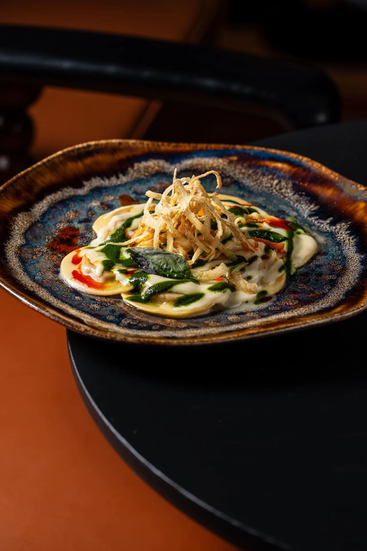 Round ravioli under cream and green herb oil, topped with a nest of crisp fried potato straws and a basil leaf, served on a blue reactive-glaze plate at GINA