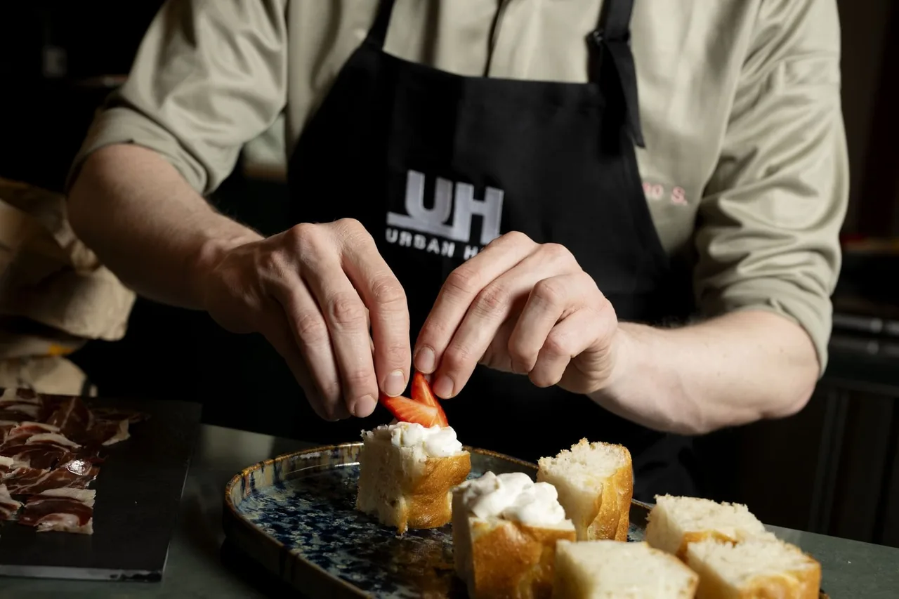 A cook in a black apron finishing squares of bread topped with whipped cheese and a sliced strawberry, a slate of sliced jamon alongside