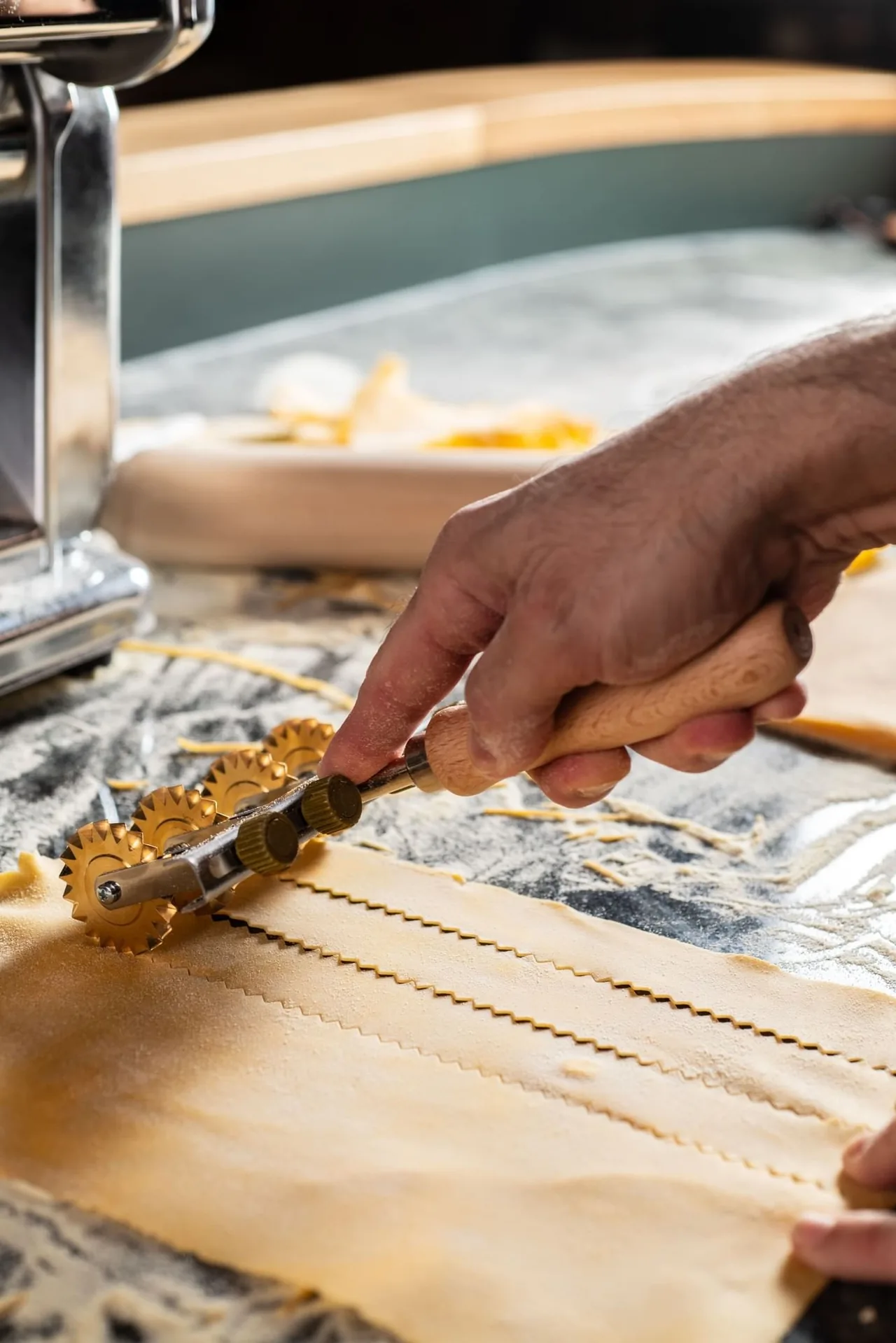 A hand running a brass multi-wheel cutter through a sheet of fresh egg pasta, the pasta machine and a flour-dusted board behind