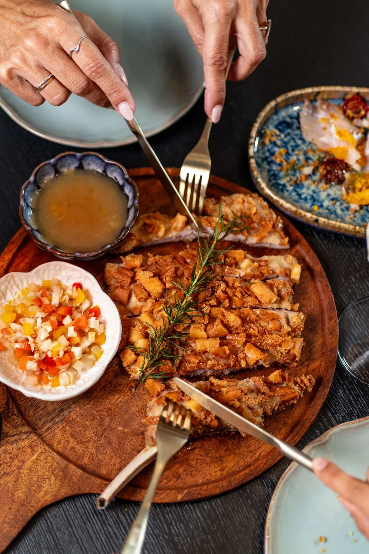 Overhead of a breaded veal cutlet sliced on a round wooden board with a rosemary sprig, a bowl of jus and a bowl of diced vegetable relish, a plate of cured fish crudo to one side, diners' hands reaching in with cutlery