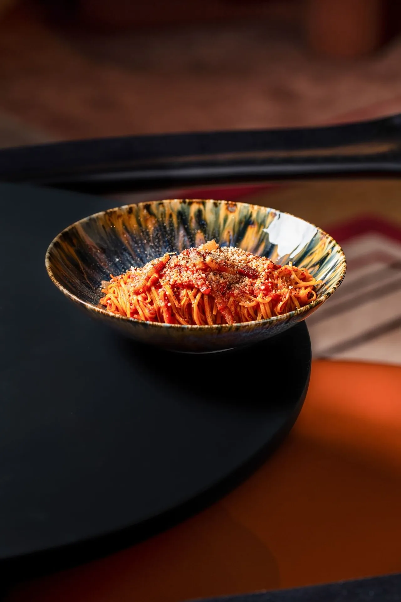 Spaghetti in tomato sauce with strips of guanciale and a heavy dusting of grated cheese, served in a mottled blue and amber glazed bowl
