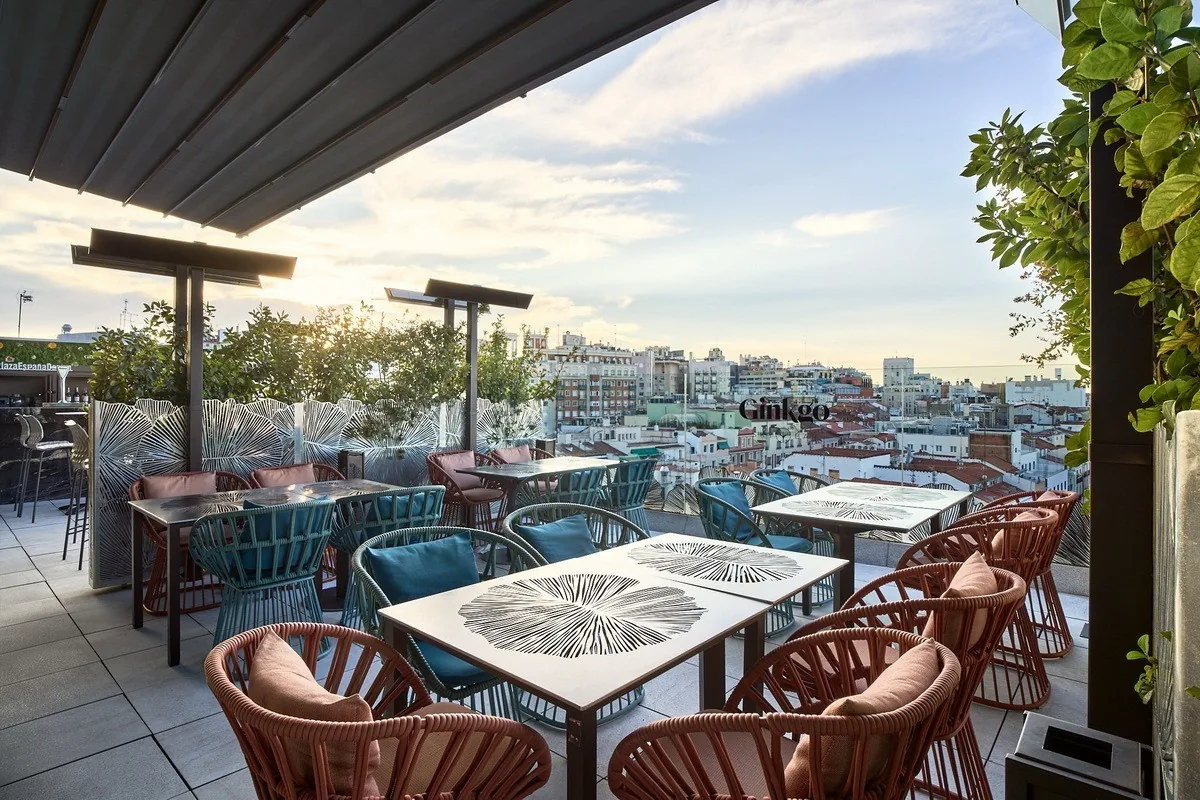 A rooftop terrace with wicker chairs and tables under an awning above the city