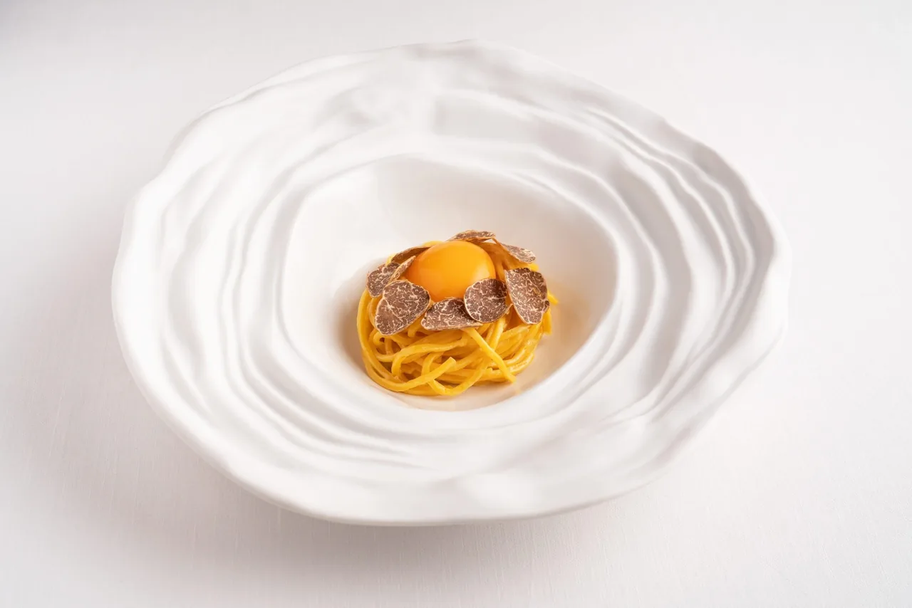 A nest of golden tagliolini topped with an egg yolk on a white rippled plate