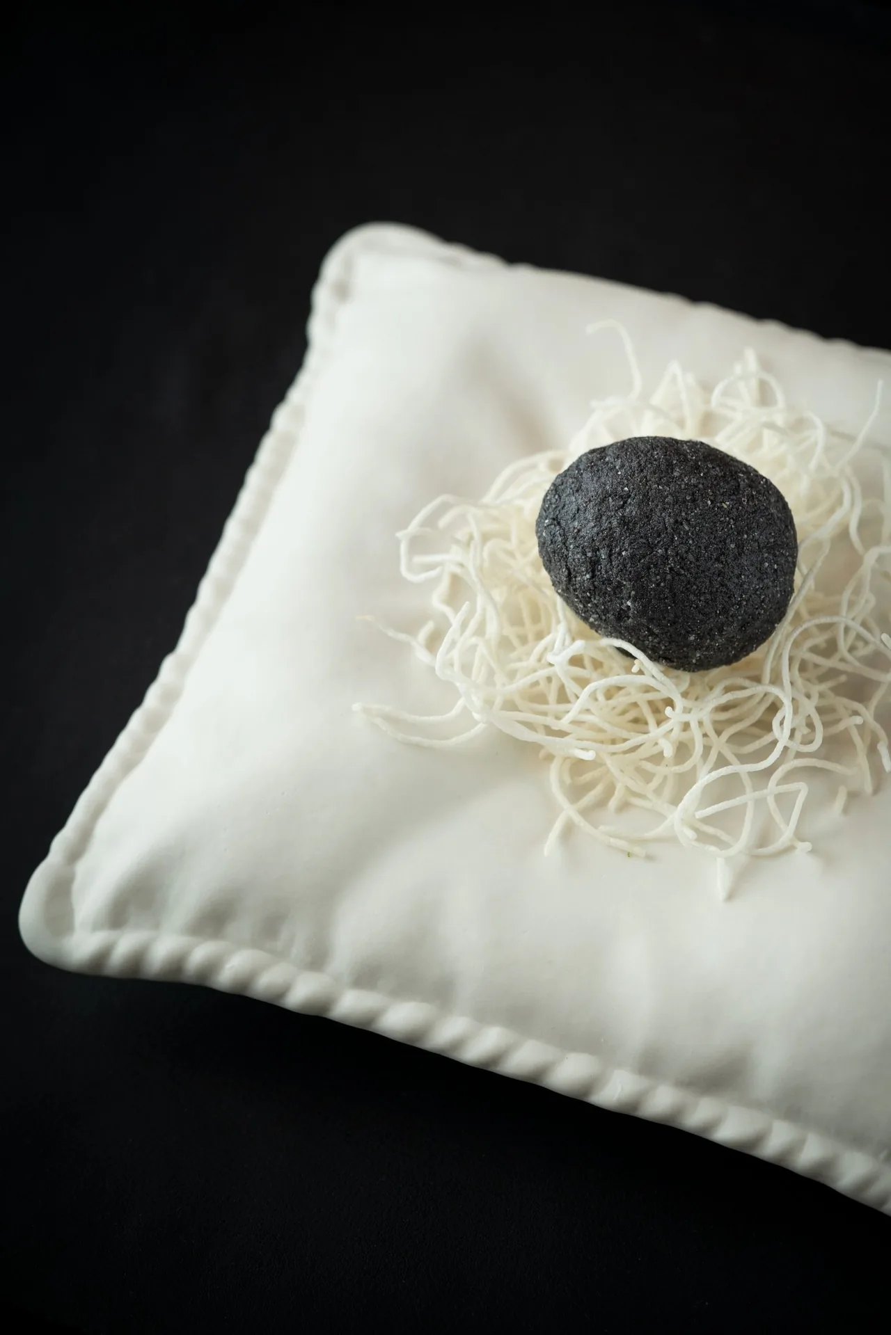 A small white cushion-shaped plate holding fine noodles topped with a dark truffle
