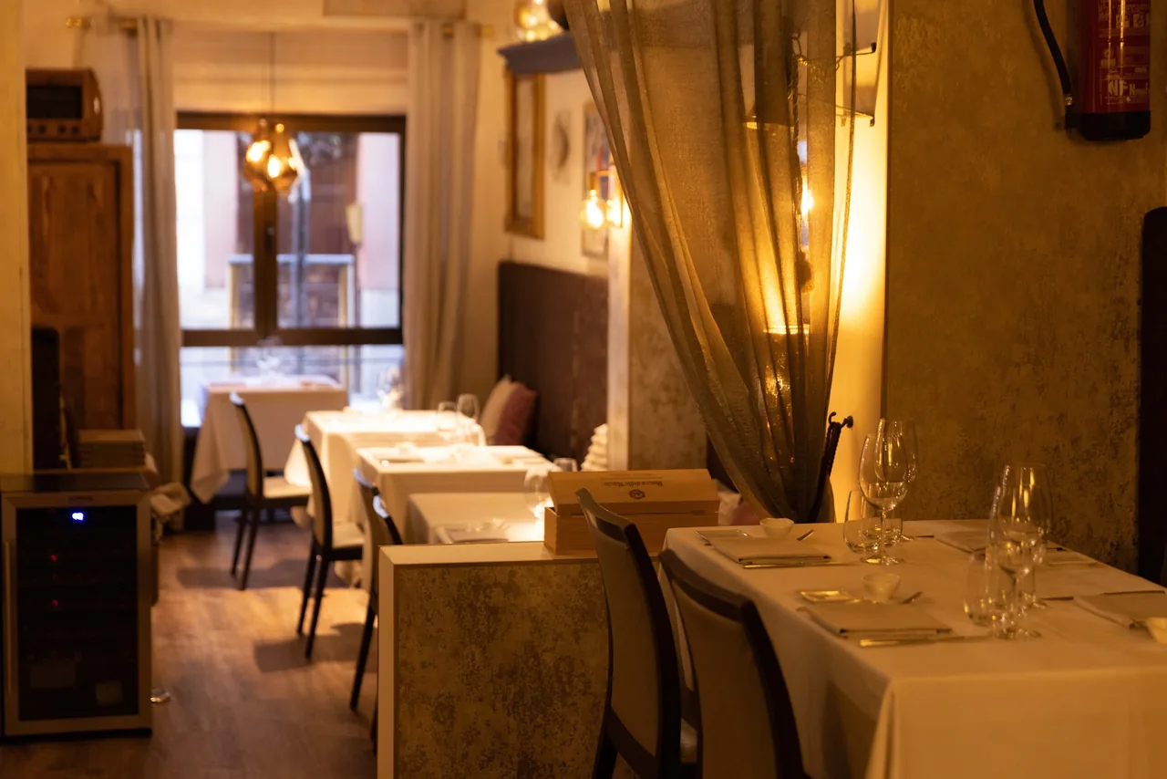 A narrow warmly lit dining room with white-clothed tables, sheer curtains and a window at the end