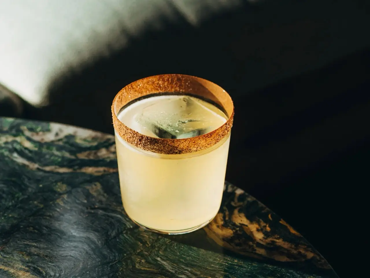 Pale yellow cocktail with a spiced sugar rim over a large ice cube in a tumbler on a green marble table