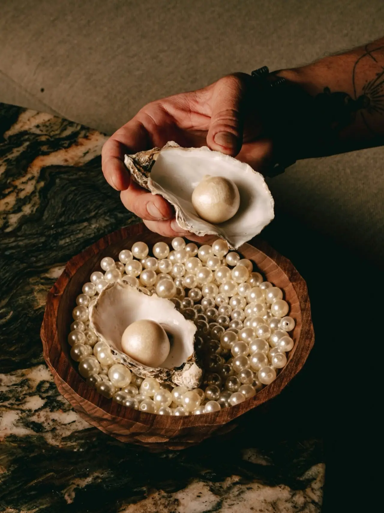 Hands presenting a pale spherical course in an oyster shell above a wooden bowl filled with pearls holding a second shell