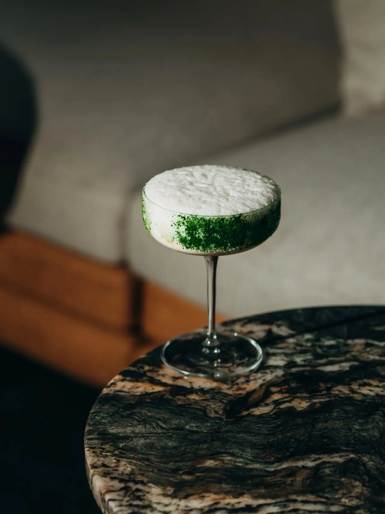 Frothy white cocktail with a green sugar rim in a stemmed coupe on a dark marble table