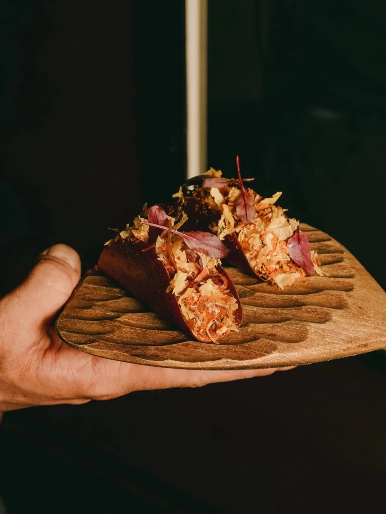 Hand holding a carved wooden board with two gourmet tacos topped with bonito flakes and red micro-herbs