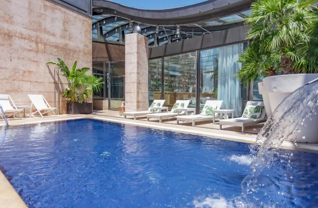 Rooftop pool terrace with white sun loungers, palm plants, a small waterfall and a glass-fronted bar behind
