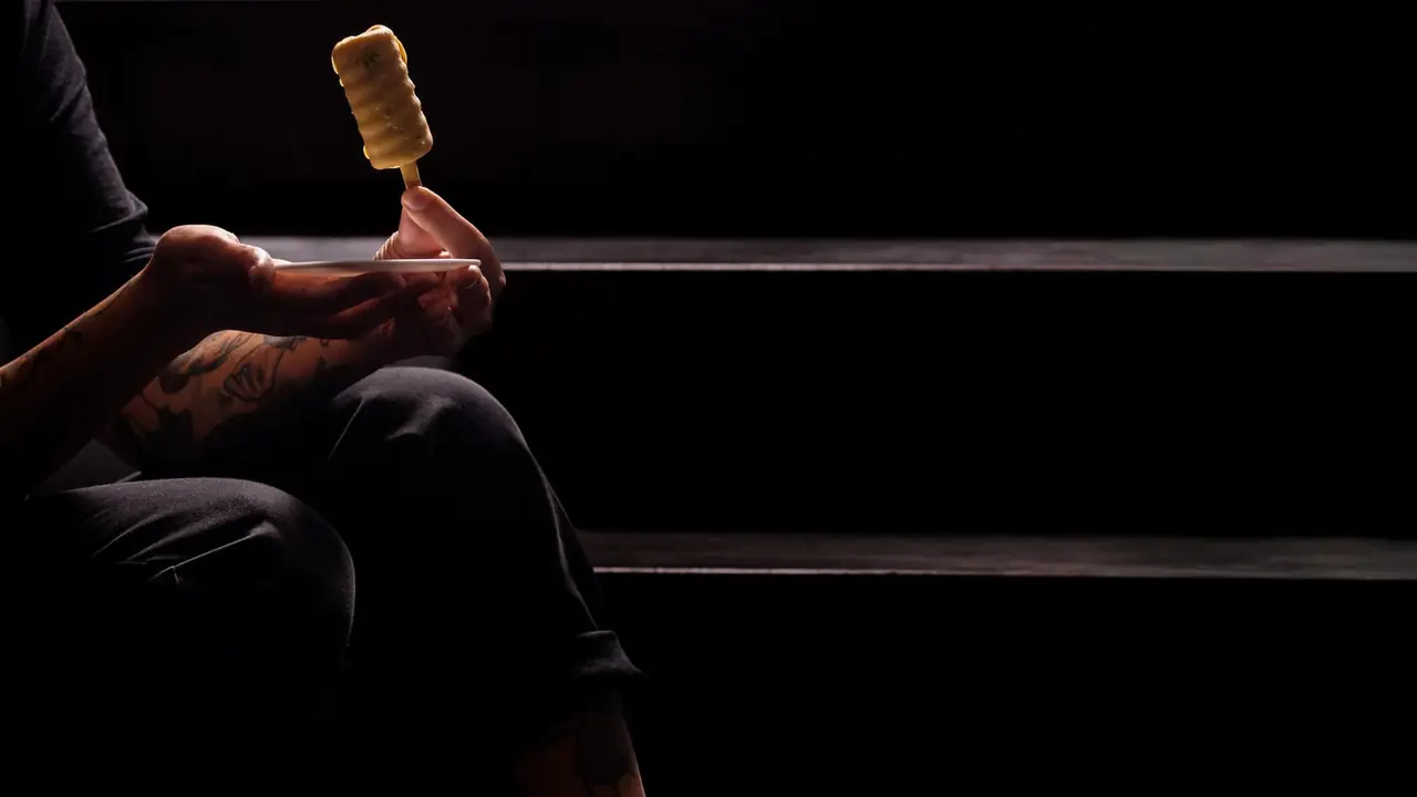 Tattooed hands holding a narrow white plate up to the light, a pale savoury lolly on a wooden stick standing upright on it, in a darkened dining room