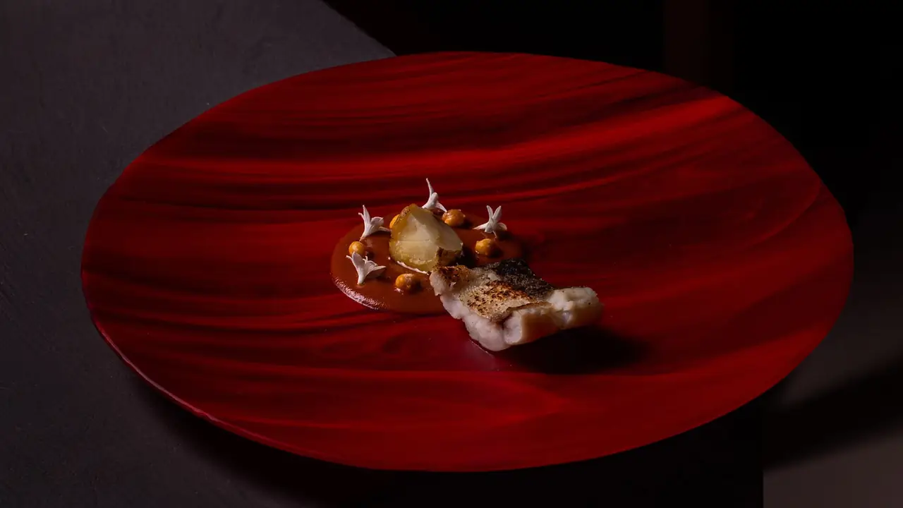 Crisp-skinned white fish fillet on a swirled red glass plate with a smear of red-brown sauce, a peeled potato, chickpeas and small white flowers