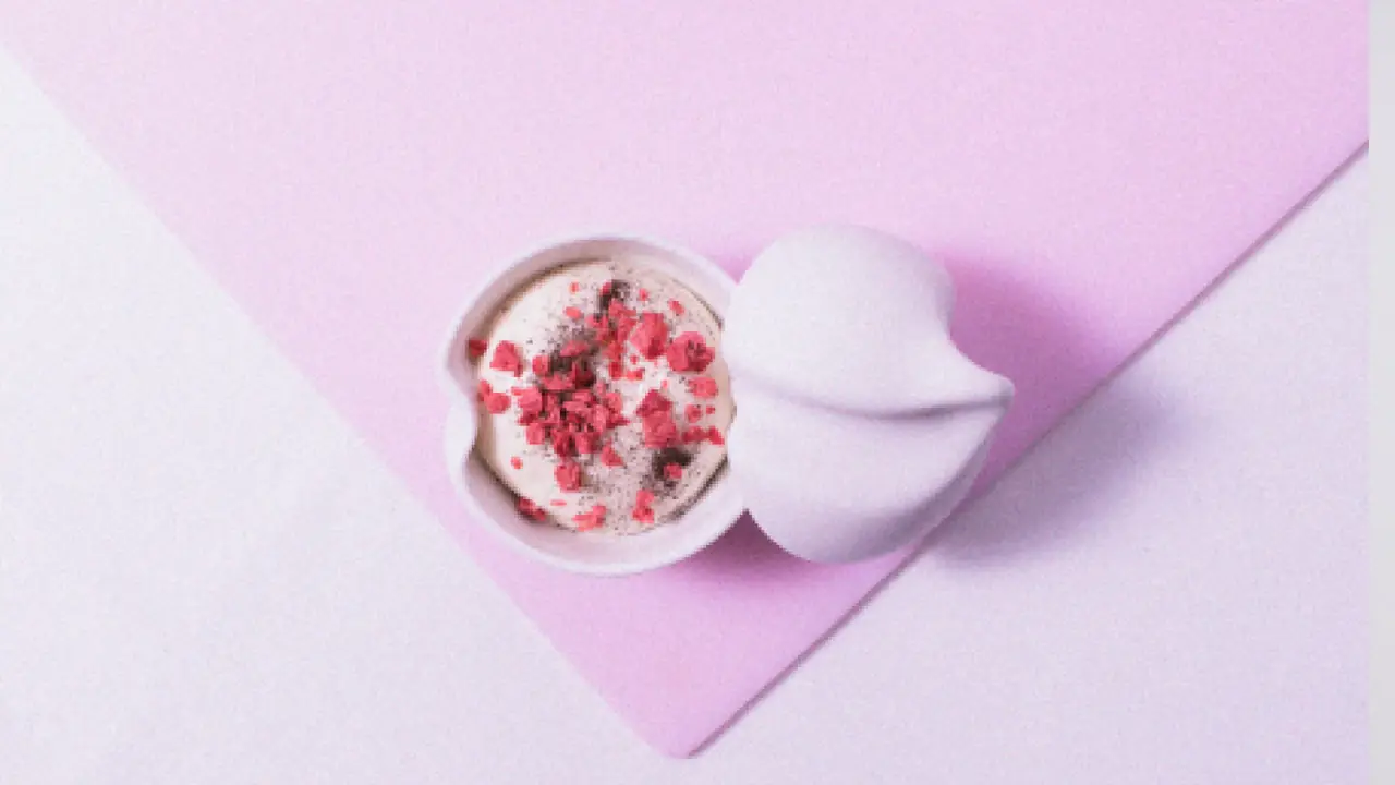 Overhead of a small white lidded ceramic pot holding pale mousse scattered with freeze-dried raspberry pieces, on folded pink paper