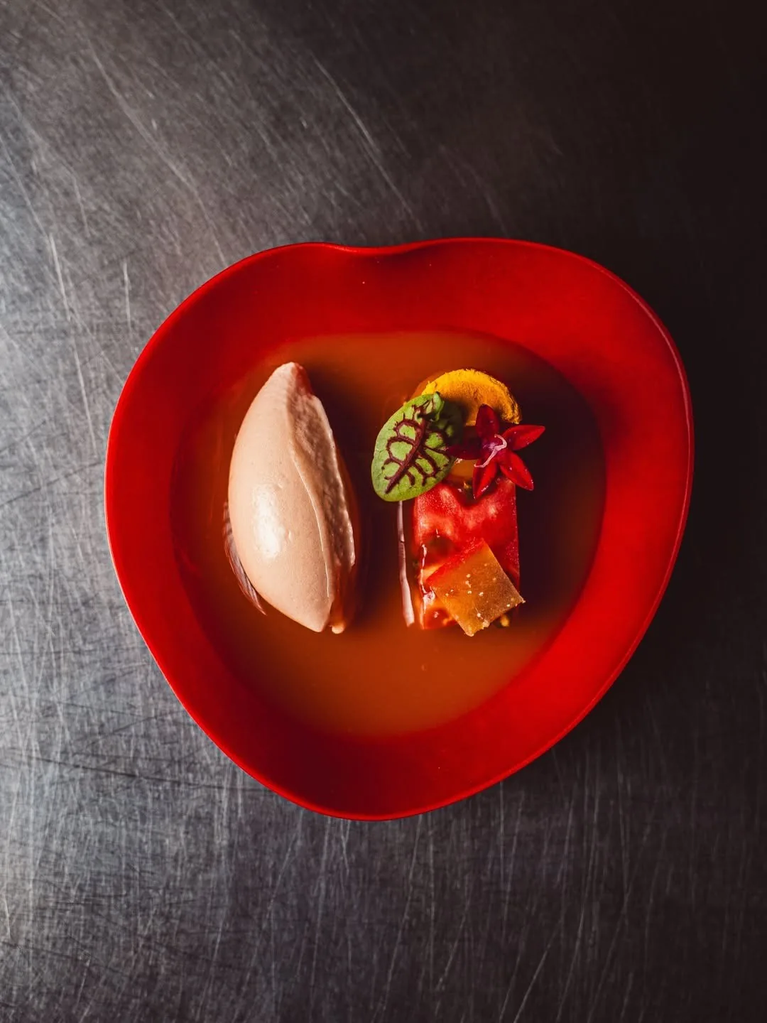 A red heart-shaped bowl with a pale scoop and colourful garnishes on a dark metal surface