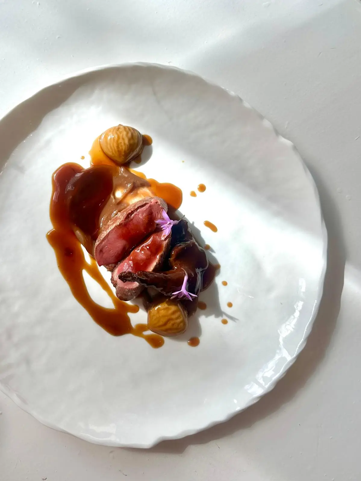 Sliced pink duck breast with jus, chestnuts and a small purple flower garnish on a white plate