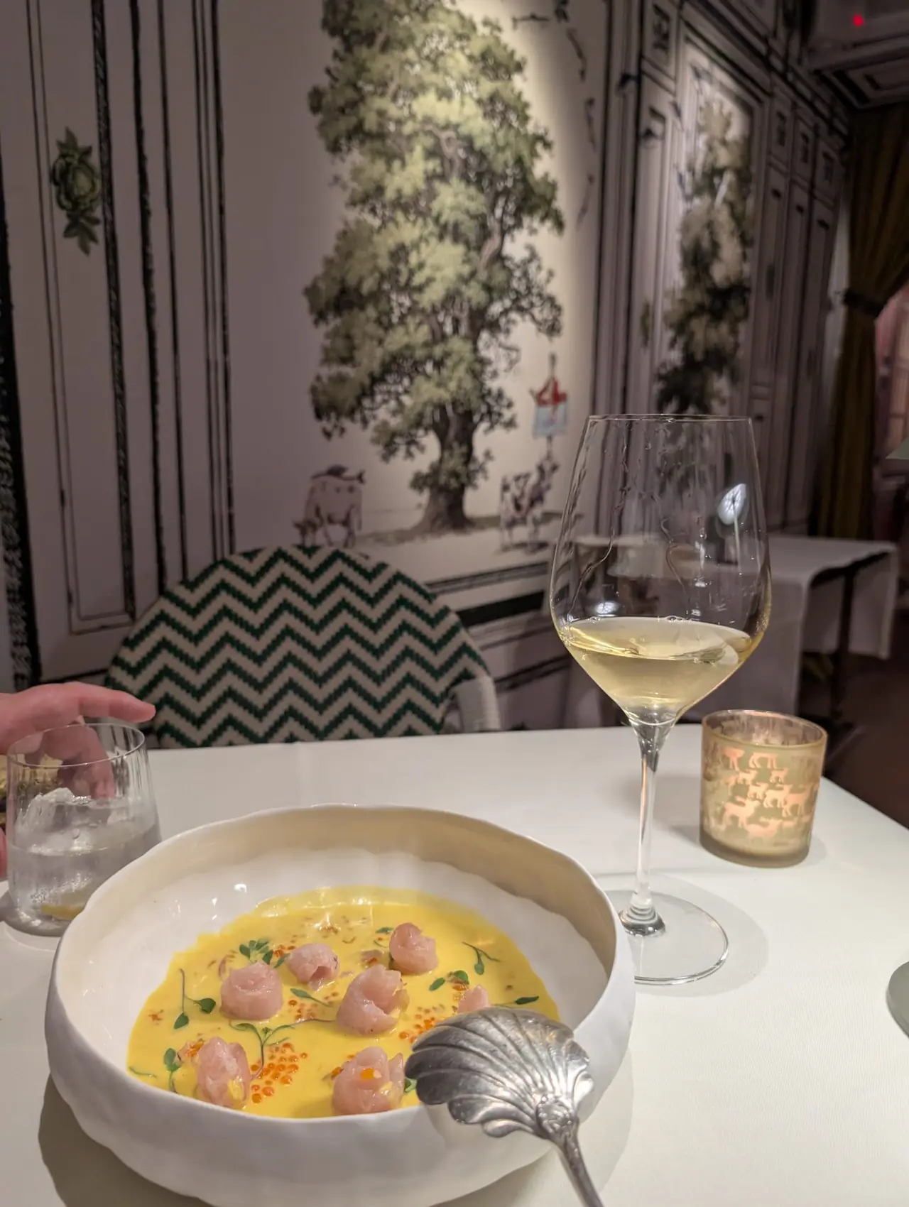 Bowl of fish tartare pieces with roe in a yellow broth, glass of white wine on the table