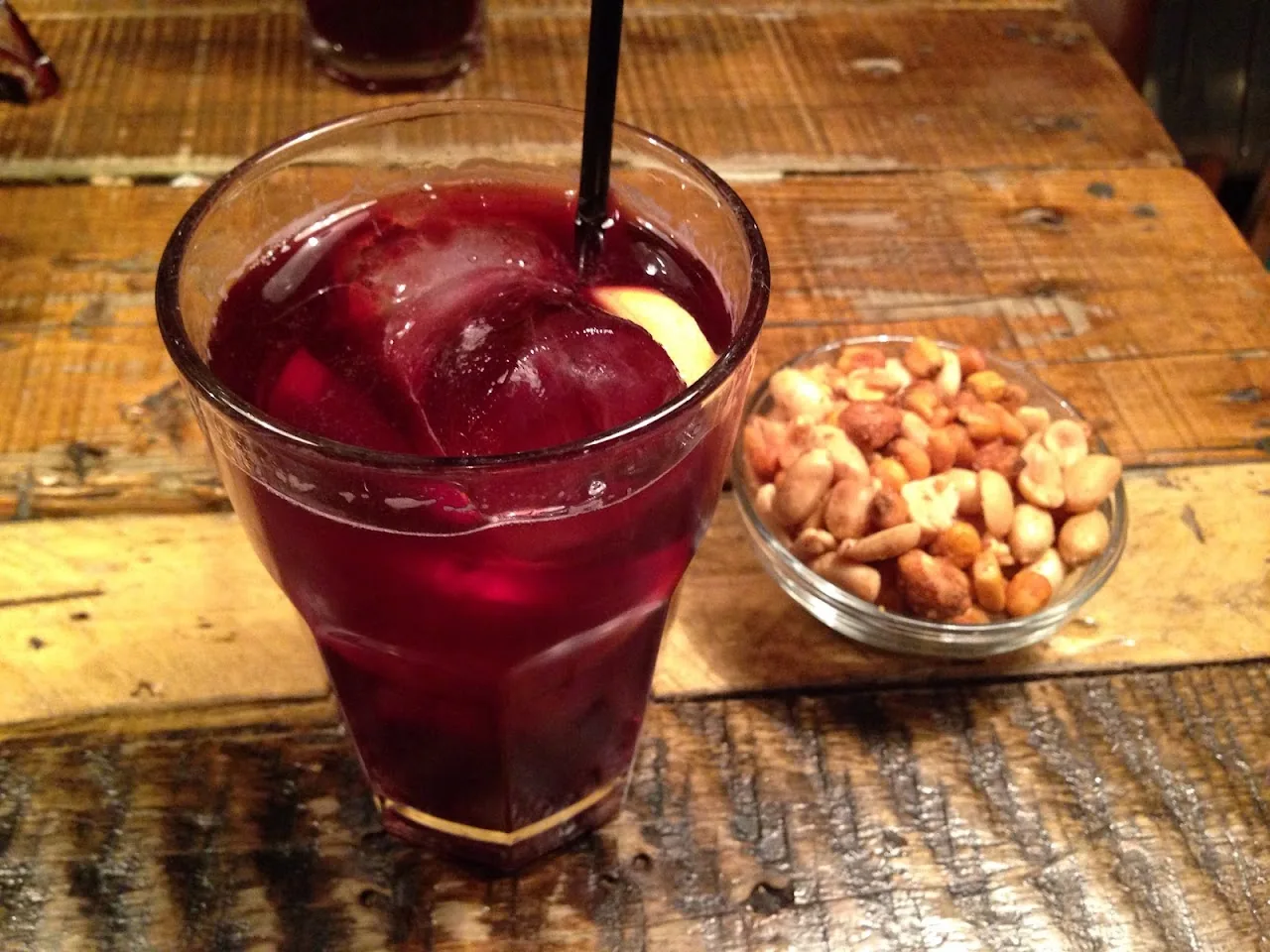 A red cocktail with ice and a straw, next to a bowl of peanuts