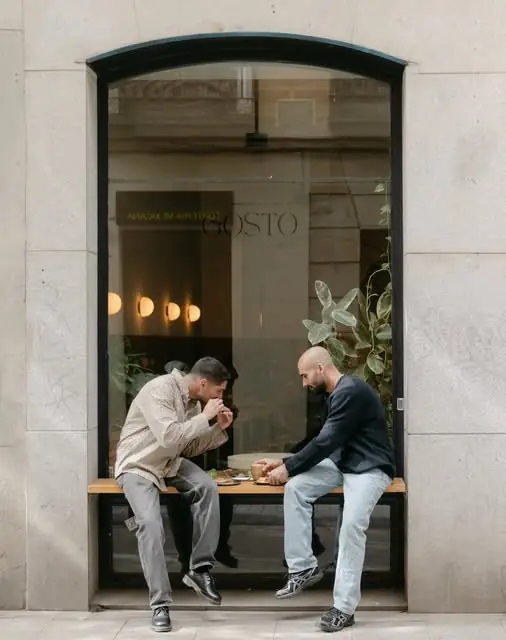 Cafe storefront with a large arched window bearing GOSTO signage, two people eating on a wooden window-ledge bench on the street