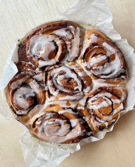 Round pull-apart batch of glazed cinnamon rolls with white icing, resting in parchment paper on a wood surface