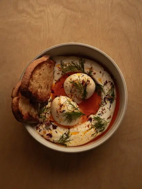 Turkish eggs, two poached eggs over whipped yogurt with chilli oil and dill, a wedge of grilled sourdough on the side, in a bowl on wood