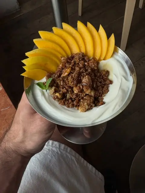 Bowl of thick yogurt topped with clustered granola, walnuts and a fan of fresh mango slices with mint, held up on a footed dish